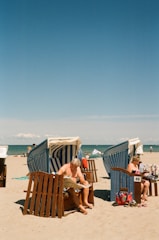A sunny beach scene with two people relaxing in wicker beach chairs. One person is reading a newspaper while another is engrossed in a book. The beach chairs have blue and white striped canopies. The sand is light and the sky is clear with a deep blue hue extending to the horizon. The ocean is calm in the background.