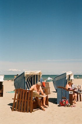 A sunny beach scene with two people relaxing in wicker beach chairs. One person is reading a newspaper while another is engrossed in a book. The beach chairs have blue and white striped canopies. The sand is light and the sky is clear with a deep blue hue extending to the horizon. The ocean is calm in the background.