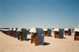 A sandy beach with multiple blue and white striped wicker beach chairs arranged in rows, facing the sea under a clear blue sky. Some chairs have white canopies, and there are few people in the distance.