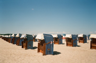 A sandy beach with multiple blue and white striped wicker beach chairs arranged in rows, facing the sea under a clear blue sky. Some chairs have white canopies, and there are few people in the distance.
