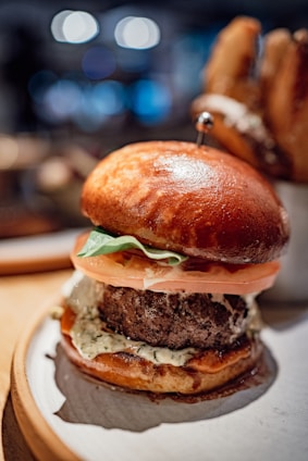A close-up of a gourmet hamburger featuring a glossy, golden-brown bun. The burger is stacked with fresh tomato slices, crisp lettuce, and a thick beef patty, adorned with a creamy sauce. The background is softly blurred, highlighting the intricate textures of the ingredients.