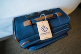 A blue travel bag with brown leather accents sits on a surface. Attached to the handle is a beige tag with the words 'FRUGAL FLYER,' featuring a stylized airplane logo and text promoting travel savings.
