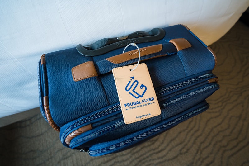 A blue travel bag with brown leather accents sits on a surface. Attached to the handle is a beige tag with the words 'FRUGAL FLYER,' featuring a stylized airplane logo and text promoting travel savings.