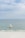 A yellow sailboat with a colorful sail featuring a logo is gliding across calm, clear ocean waters. The background shows a vast blue sky with scattered clouds, and a beach with white sand is visible in the foreground.