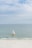 A yellow sailboat with a colorful sail featuring a logo is gliding across calm, clear ocean waters. The background shows a vast blue sky with scattered clouds, and a beach with white sand is visible in the foreground.
