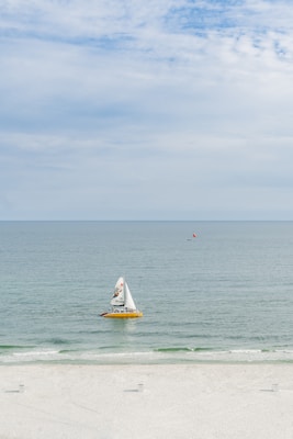 A yellow sailboat with a colorful sail featuring a logo is gliding across calm, clear ocean waters. The background shows a vast blue sky with scattered clouds, and a beach with white sand is visible in the foreground.