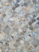 An intricate mosaic pattern composed of irregularly shaped tiles featuring a mix of designs. The designs vary from floral motifs to geometric shapes, with a color palette of blues, grays, whites, and browns. The surface texture appears rough and uneven, providing a visually appealing and tactile experience.