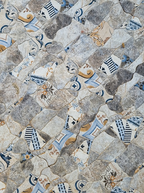 An intricate mosaic pattern composed of irregularly shaped tiles featuring a mix of designs. The designs vary from floral motifs to geometric shapes, with a color palette of blues, grays, whites, and browns. The surface texture appears rough and uneven, providing a visually appealing and tactile experience.