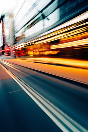 A dynamic city street scene showing motion blur from fast-moving lights, suggesting cars or other vehicles. Buildings with illuminated windows and various light sources create a vivid and energetic atmosphere.