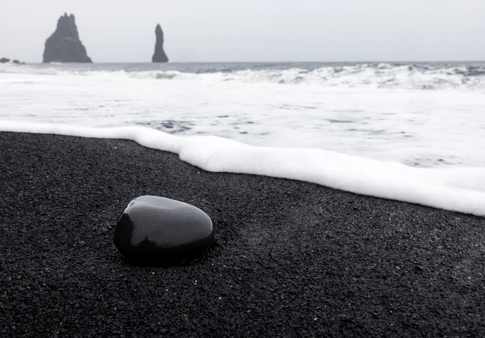 The image depicts a black and white scene at the beach, with two large rock formations in the background. The rocks are situated on the left side of the frame, standing tall against the backdrop of the ocean. The water is a light blue color, contrasting with the darker tones of the rocks. In the foreground, there's a small pebble resting on the dark sand near the center of the image.