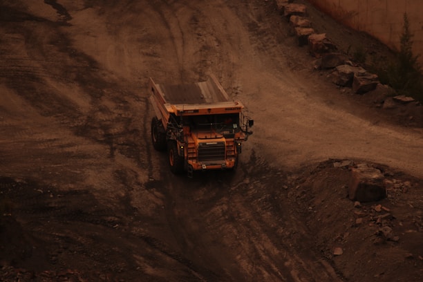 A large dump truck is navigating through a rugged terrain pathway surrounded by rocks and dirt. The environment appears industrial, possibly a mining site with earthy colors dominating the scene.