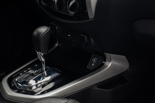 An interior view of a car's dashboard featuring an automatic gear shift lever. The gear shift is metallic with a black handle, and the panel includes a USB port. Surrounding controls for the vehicle's air conditioning system and power outlets can be seen.