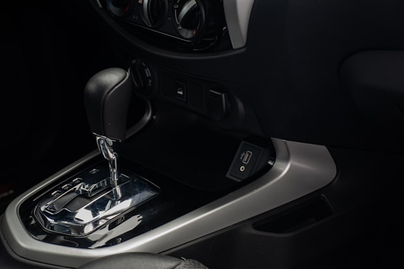 An interior view of a car's dashboard featuring an automatic gear shift lever. The gear shift is metallic with a black handle, and the panel includes a USB port. Surrounding controls for the vehicle's air conditioning system and power outlets can be seen.