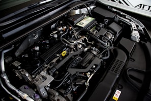 A detailed view of a car engine compartment, showcasing various components such as hoses, belts, and a battery. The metallic parts and wires are neatly arranged, with some labeling visible on parts. The lighting emphasizes the mechanical complexity and precision of the engine design.