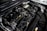 A detailed view of a car engine compartment, showcasing various components such as hoses, belts, and a battery. The metallic parts and wires are neatly arranged, with some labeling visible on parts. The lighting emphasizes the mechanical complexity and precision of the engine design.
