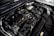 A detailed view of a car engine compartment, showcasing various components such as hoses, belts, and a battery. The metallic parts and wires are neatly arranged, with some labeling visible on parts. The lighting emphasizes the mechanical complexity and precision of the engine design.