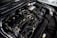 A detailed view of a car engine compartment, showcasing various components such as hoses, belts, and a battery. The metallic parts and wires are neatly arranged, with some labeling visible on parts. The lighting emphasizes the mechanical complexity and precision of the engine design.