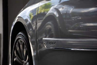 Close-up of a car's side, focusing on the wheel and a chrome emblem with the word 'Corolla.' The car surface has a glossy finish, reflecting light and the surrounding environment.