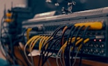 A close-up view of a complex audio or video equipment setup with numerous cables of different colors connected to various ports on a rack-mounted device. The cables are organized in multiple rows, with visible plugs, and some indicator lights are lit on the equipment.