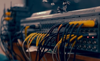 A close-up view of a complex audio or video equipment setup with numerous cables of different colors connected to various ports on a rack-mounted device. The cables are organized in multiple rows, with visible plugs, and some indicator lights are lit on the equipment.