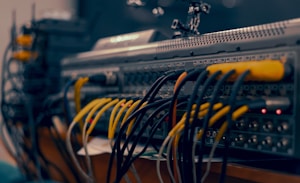 A close-up view of a complex audio or video equipment setup with numerous cables of different colors connected to various ports on a rack-mounted device. The cables are organized in multiple rows, with visible plugs, and some indicator lights are lit on the equipment.