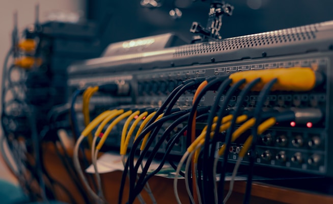 A close-up view of a complex audio or video equipment setup with numerous cables of different colors connected to various ports on a rack-mounted device. The cables are organized in multiple rows, with visible plugs, and some indicator lights are lit on the equipment.