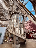 A detailed view of an ornate iron and glass elevator structure within an elaborate metal framework. The elevator is enclosed in glass panels, surrounded by a complex lattice of beams and girders, reflecting industrial architecture. Sunlight casts shadows and highlights various structures, and an open sky is visible through some parts.