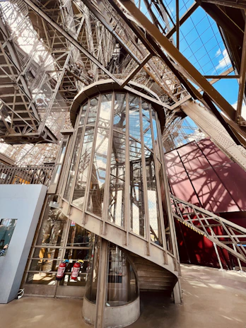 A detailed view of an ornate iron and glass elevator structure within an elaborate metal framework. The elevator is enclosed in glass panels, surrounded by a complex lattice of beams and girders, reflecting industrial architecture. Sunlight casts shadows and highlights various structures, and an open sky is visible through some parts.