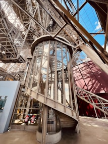 A detailed view of an ornate iron and glass elevator structure within an elaborate metal framework. The elevator is enclosed in glass panels, surrounded by a complex lattice of beams and girders, reflecting industrial architecture. Sunlight casts shadows and highlights various structures, and an open sky is visible through some parts.