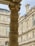 A detailed stone column with intricate carvings stands prominently against the backdrop of classic architectural buildings. The facade features multiple windows and elaborate decorative elements, showcasing historical and artistic craftsmanship.