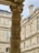 A detailed stone column with intricate carvings stands prominently against the backdrop of classic architectural buildings. The facade features multiple windows and elaborate decorative elements, showcasing historical and artistic craftsmanship.