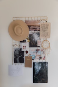 A mood board featuring a variety of items hanging on a metal grid. These items include a beige hat, a dreamcatcher, black and white photos, landscape images, notes with handwritten text, and an envelope labeled 'NOTES'. The arrangement conveys a sense of nostalgia and inspiration.
