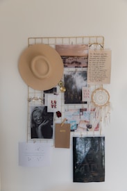 A mood board featuring a variety of items hanging on a metal grid. These items include a beige hat, a dreamcatcher, black and white photos, landscape images, notes with handwritten text, and an envelope labeled 'NOTES'. The arrangement conveys a sense of nostalgia and inspiration.