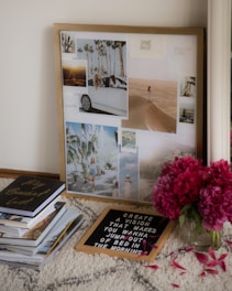 A collage of photographs in a frame is leaning against a wall. The images include a desert scene, palm trees, and someone leaning out of a car. Nearby is a stack of books with 'My Bucket List' on the top, a letter board with motivational text, and a vase of bright pink flowers.