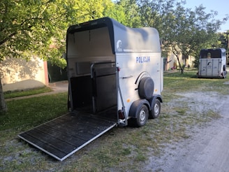 A horse trailer with the word 'POLICJA' on the side is parked on a grassy area, near a tree casting shadows. Another trailer is visible in the background. The trailers are designed with a ramp for loading and unloading horses.