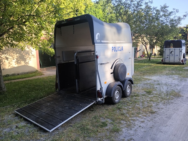 A horse trailer with the word 'POLICJA' on the side is parked on a grassy area, near a tree casting shadows. Another trailer is visible in the background. The trailers are designed with a ramp for loading and unloading horses.