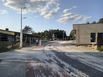 A newly constructed road with interlocking bricks and surrounding buildings under construction. Two people are walking side by side along the road. The sky is clear with scattered clouds, and there are trees in the background.
