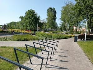 A park scene with a path leading through well-maintained grassy areas and organized flower beds. Several people are relaxing on benches near a small fountain. Trees provide shade, and there is a children's playground visible in the background.