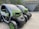 Two compact electric vehicles are parked in an outdoor setting, featuring green and black coloring. The vehicles have a futuristic design with low-profile wheels and transparent roofs. They are situated near a building with a grassy roof, and a scenic background of green hills and trees.