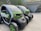 Two compact electric vehicles are parked in an outdoor setting, featuring green and black coloring. The vehicles have a futuristic design with low-profile wheels and transparent roofs. They are situated near a building with a grassy roof, and a scenic background of green hills and trees.