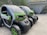 Two compact electric vehicles are parked in an outdoor setting, featuring green and black coloring. The vehicles have a futuristic design with low-profile wheels and transparent roofs. They are situated near a building with a grassy roof, and a scenic background of green hills and trees.