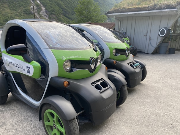 Two compact electric vehicles are parked in an outdoor setting, featuring green and black coloring. The vehicles have a futuristic design with low-profile wheels and transparent roofs. They are situated near a building with a grassy roof, and a scenic background of green hills and trees.