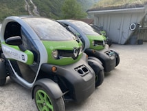 Two compact electric vehicles are parked in an outdoor setting, featuring green and black coloring. The vehicles have a futuristic design with low-profile wheels and transparent roofs. They are situated near a building with a grassy roof, and a scenic background of green hills and trees.