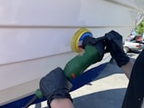 A person wearing black gloves uses a green power tool with a yellow and blue buffing pad to polish the white surface of a boat. The setting appears to be outdoors, likely in a boatyard, with a parked car visible in the background.