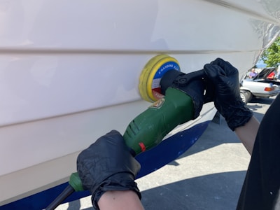 A person wearing black gloves uses a green power tool with a yellow and blue buffing pad to polish the white surface of a boat. The setting appears to be outdoors, likely in a boatyard, with a parked car visible in the background.