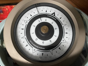 A close-up view of a gyroscope compass with a circular dial, labeled with numbers and cardinal directions. The compass has a metallic rim and a needle pointing toward the north. The background contains a blurred red object.