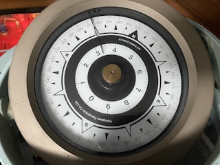 A close-up view of a gyroscope compass with a circular dial, labeled with numbers and cardinal directions. The compass has a metallic rim and a needle pointing toward the north. The background contains a blurred red object.