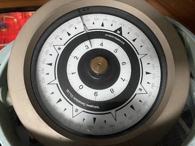 A close-up view of a gyroscope compass with a circular dial, labeled with numbers and cardinal directions. The compass has a metallic rim and a needle pointing toward the north. The background contains a blurred red object.