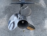 A shiny, metallic boat propeller with three blades is positioned against a textured, rough surface, possibly concrete or asphalt. The propeller has a reflective finish, displaying slight distortions of surrounding objects.