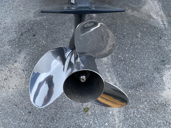 A shiny, metallic boat propeller with three blades is positioned against a textured, rough surface, possibly concrete or asphalt. The propeller has a reflective finish, displaying slight distortions of surrounding objects.